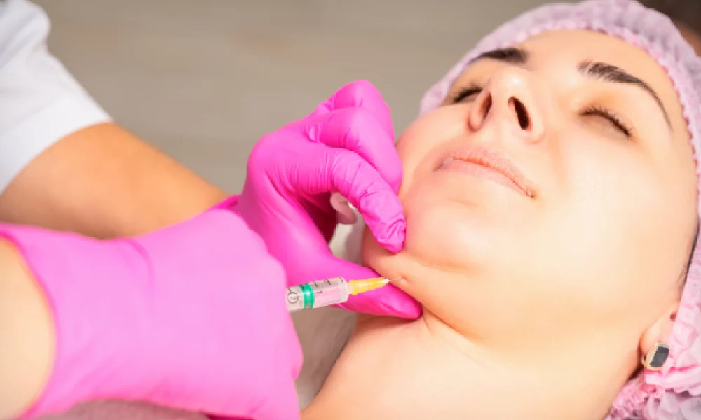 A woman receiving a Kybella injection in the chin area for double chin reduction by a medical professional wearing pink gloves in a clinical setting in Nampa, ID